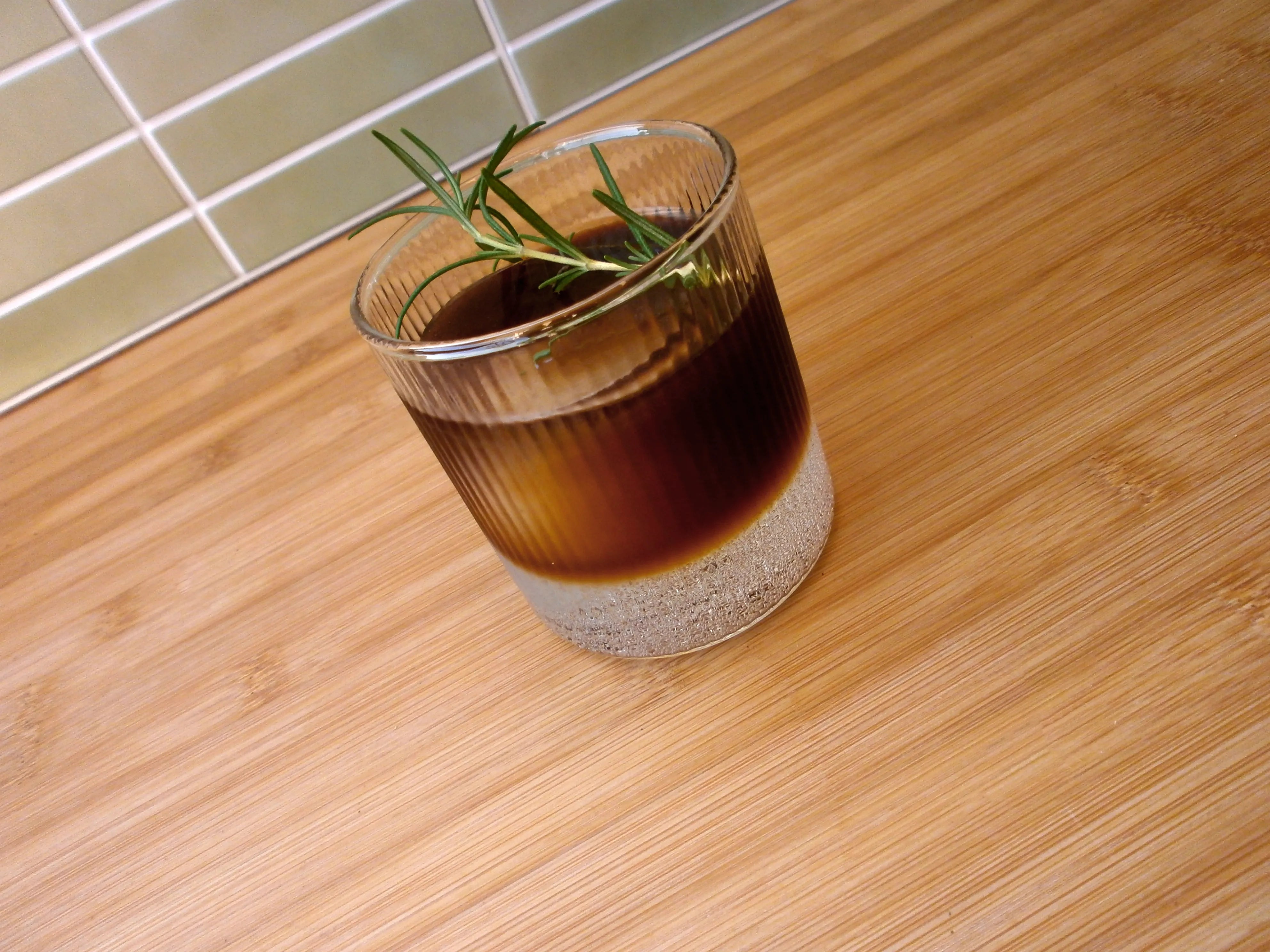 An image of a glass cup on a wooden benchtop. The cup has 3 parts tonic water and 1 part Ratio cold brew coffee tonic. It is garnished with a sprig of rosemary.
