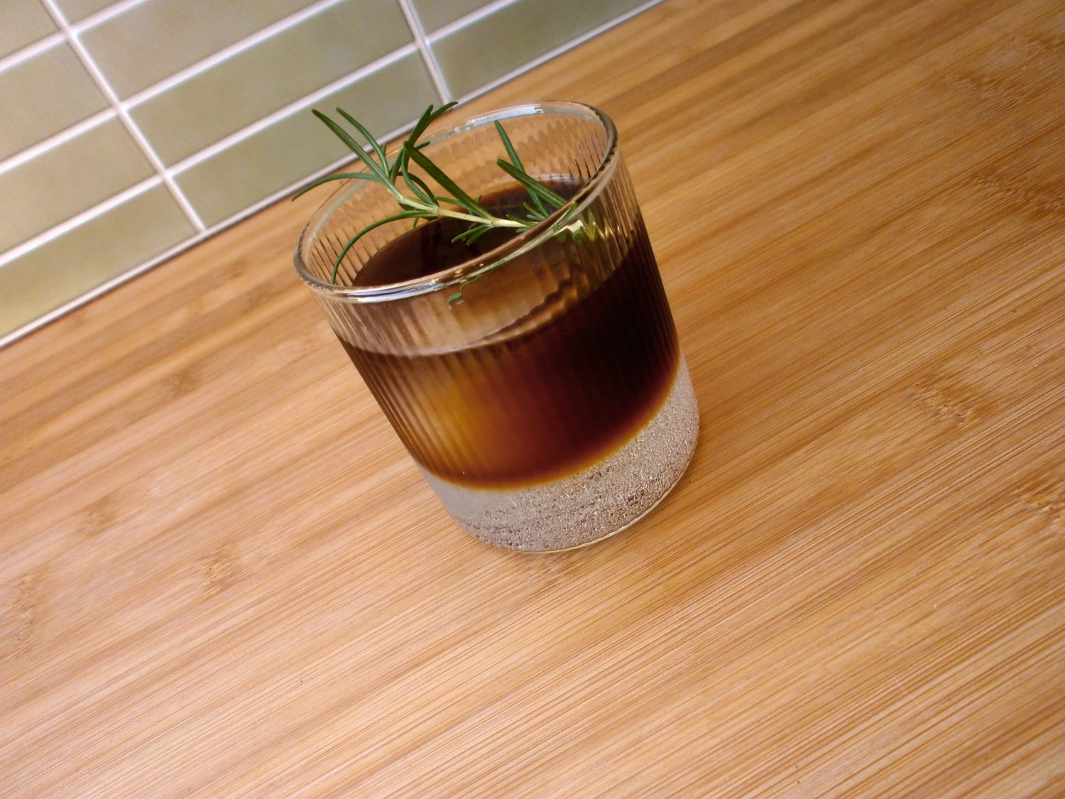 An image of a glass cup on a wooden benchtop. The cup has 3 parts tonic water and 1 part Ratio cold brew coffee tonic. It is garnished with a sprig of rosemary.