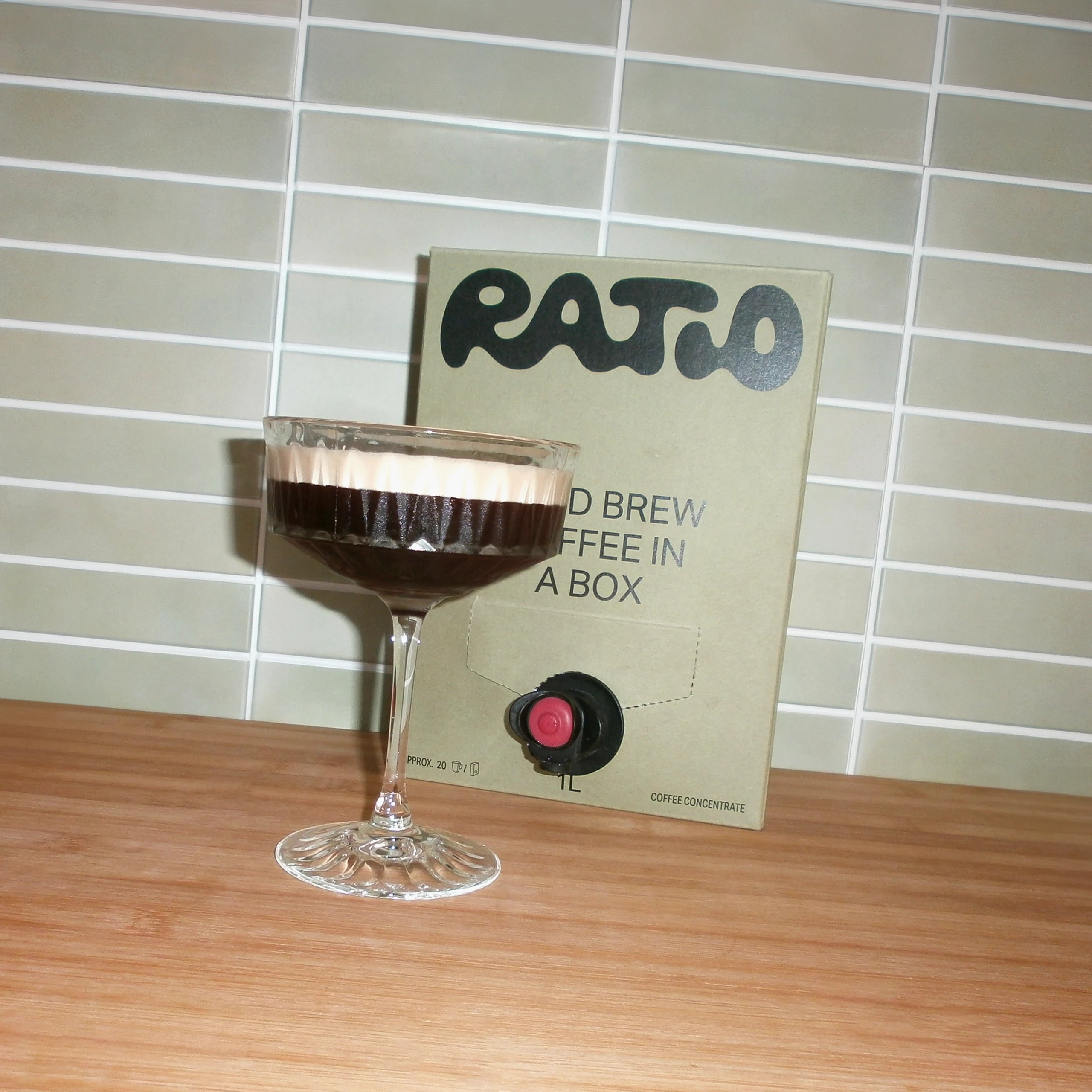 An image of a martini glass on a wooden benchtop. There is a box of Ratio cold brew coffee concentrate behind the glass. The glass contains a cold brew coffee martini.